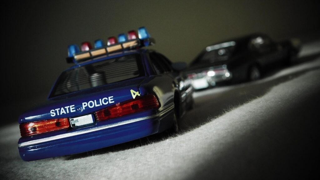 Achter op de blauwe politiewagen staat in witte letters het woord 'Police'. In de verte staat een grijze auto. Beide auto's lijken op een zachte ondergrond te staan, alsof het speelgoedauto's zijn.