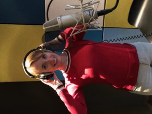Een professionele opname-studio met een grote microfoon en daarachter Susanne in een rood truitje en een grijze broek met natuurlijk een koptelefoon op haar hoofd.