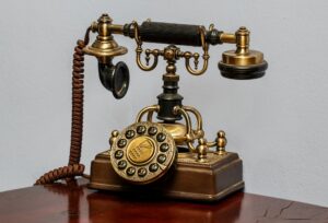 Een ouderwetse telefoon met een koperen draaischijf en een hoorn die op een sierlijke metalen haak rust. De hoorn heeft aan de ene kant een rond uiteinde en aan de andere kant een zwarte hoorn om door te praten.